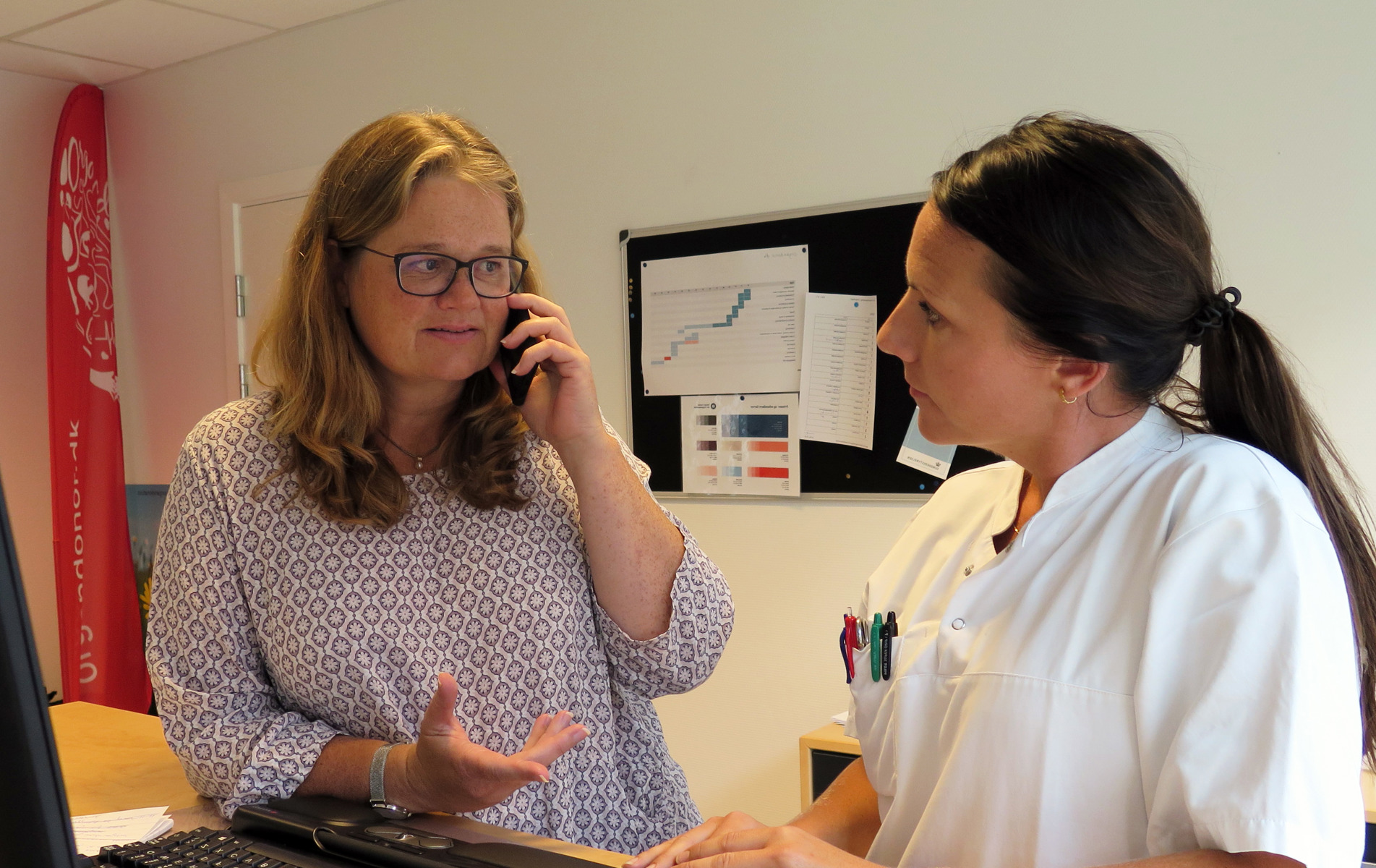 Kvinde taler i telefon, sygeplejerske står ved siden af og deltager i samtalen